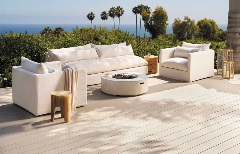 Light-colored decking boards create a clean, modern outdoor living space with white cushioned seating and a round fire pit. The deck overlooks a coastal landscape with palm trees and ocean views, highlighting a bright, low-maintenance deck surface ideal for outdoor entertaining.