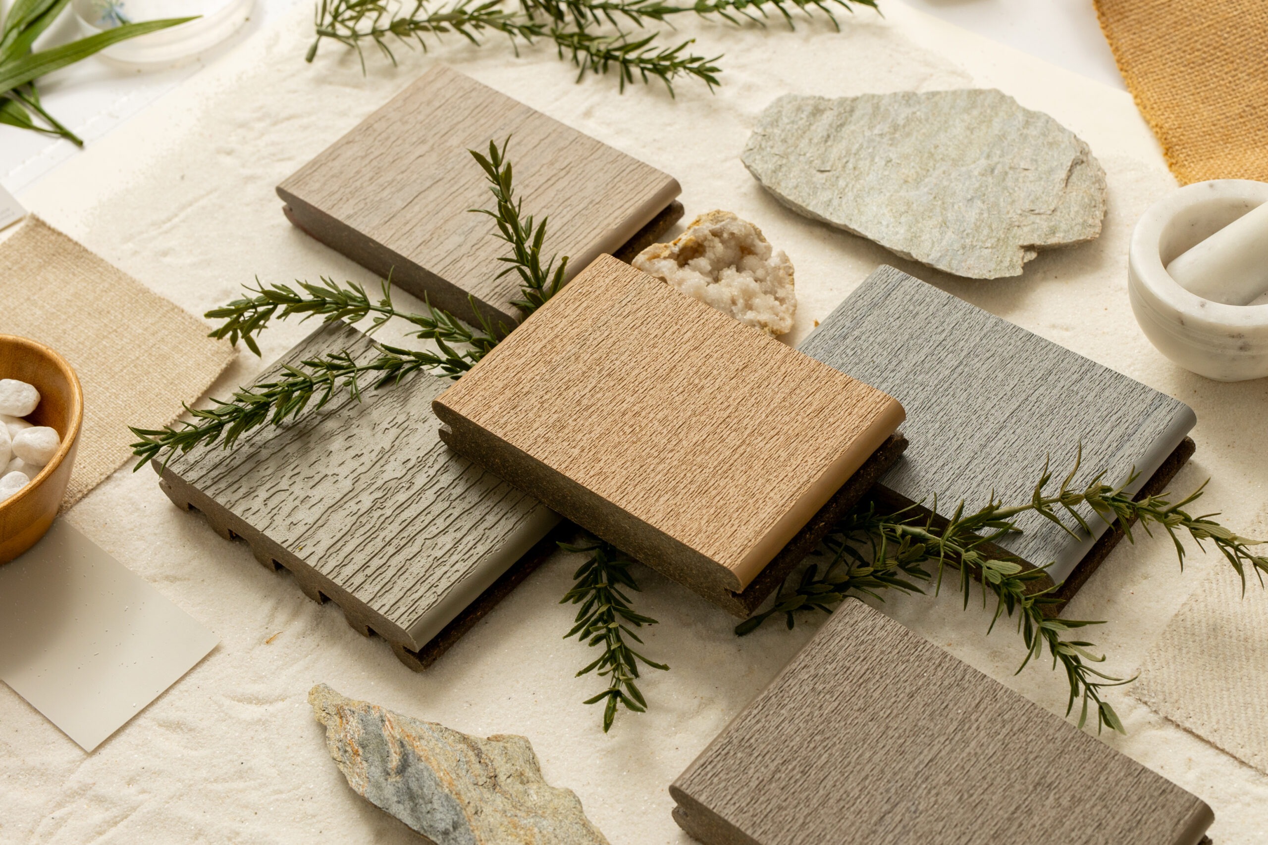 Composite decking samples in muted neutral tones displayed on a soft beige surface alongside greenery, stones, and fabric swatches. The setting evokes natural design inspiration while showcasing the subtle woodgrain and finish differences between board types. This image emphasizes how Trex deck boards can complement organic, outdoor-inspired designs.