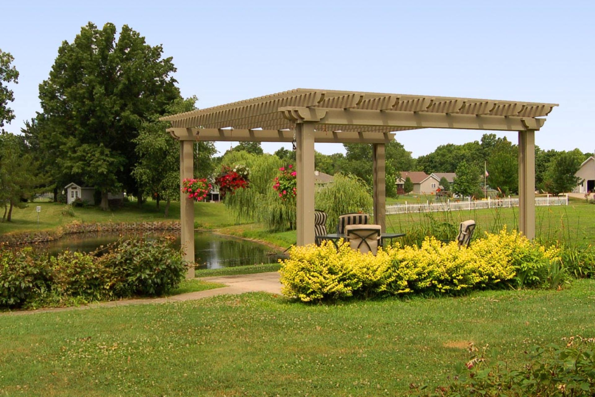Beige freestanding pergola with an open lattice roof, hanging flower baskets, and outdoor seating beside a backyard pond.