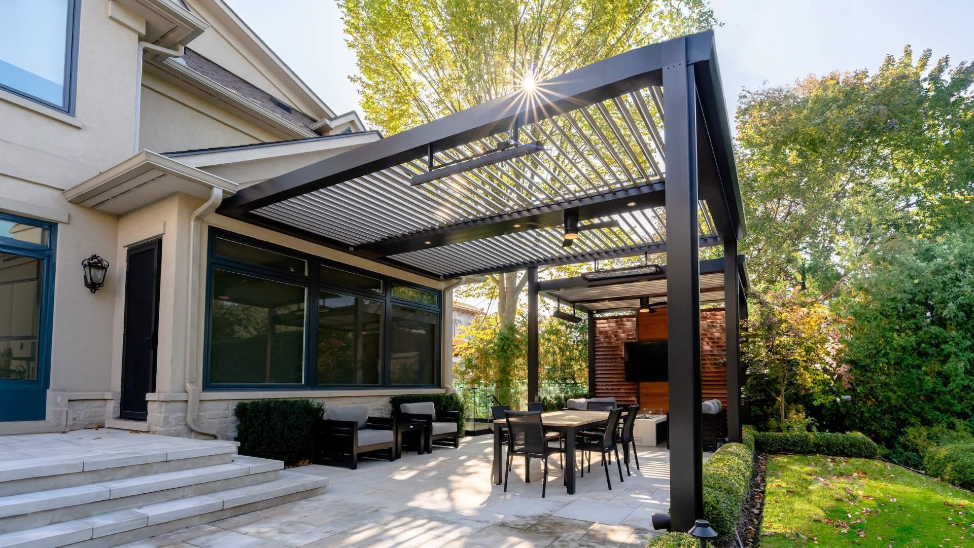 Modern black aluminum louvered pergola beside a house with outdoor dining furniture and integrated lighting.
