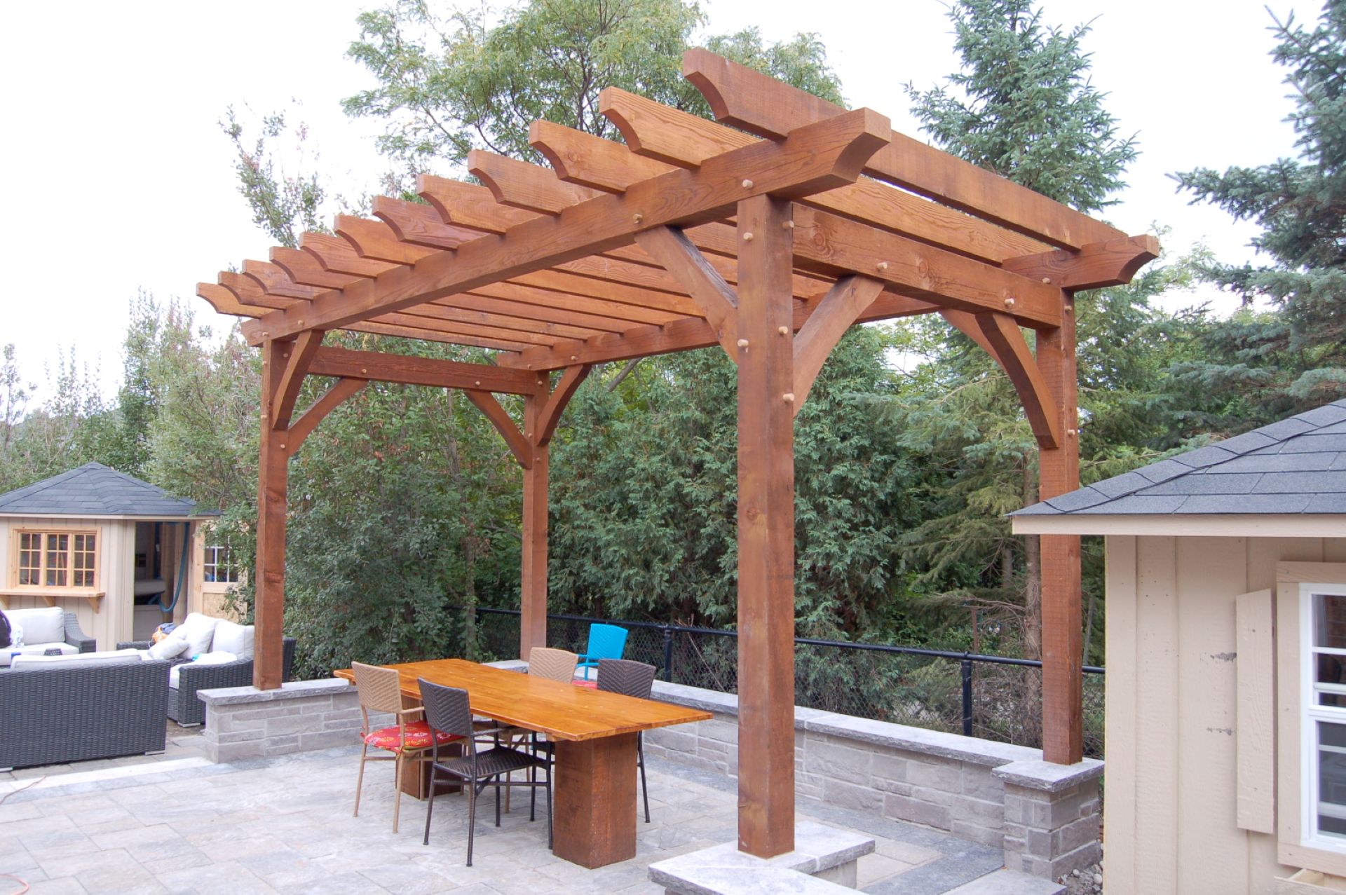 Traditional freestanding wood pergola with open rafters and decorative braces over a stone patio dining area.
