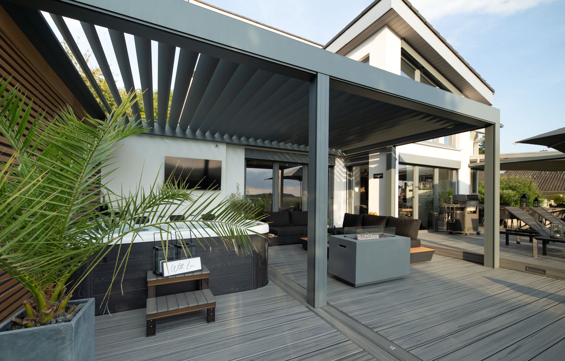 Modern gray aluminum louvered pergola over a composite deck with outdoor seating, a fire table, and a hot tub.