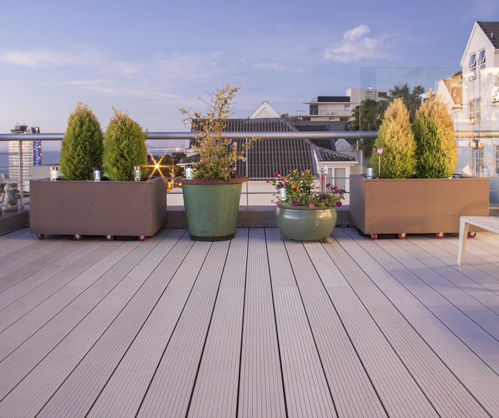 Xavia WYDE Eva-Tech decking shown on a modern rooftop patio with potted plants, overlooking a coastal cityscape at dusk.