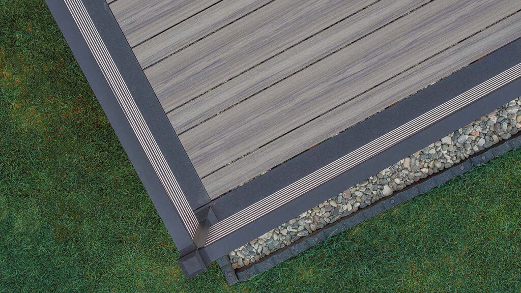 Top-down view of a deck corner featuring Deckorators Picture Frame board in Dark Slate. The frame outlines lighter-toned deck boards with clean, precise edges above decorative river rocks and vibrant green grass.