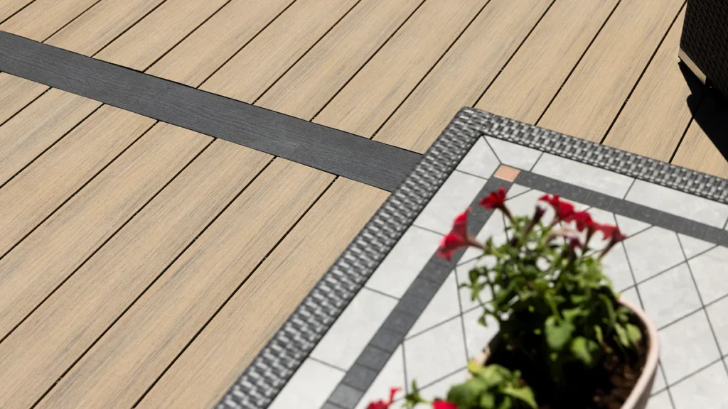 Close-up of a light wood-tone deck bordered by Deckorators Picture Frame board in Dark Slate, running horizontally across the deck as a contrasting accent beside a mosaic outdoor table and red flowers.Close-up of a light wood-tone deck bordered by Deckorators Picture Frame board in Dark Slate, running horizontally across the deck as a contrasting accent beside a mosaic outdoor table and red flowers.