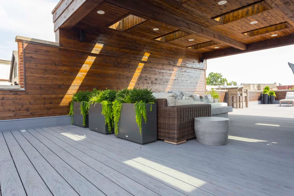 Modern rooftop deck featuring Deckorators Vault Dusk decking with warm wood accents, wicker seating, and lush green planters under a wooden pergola.