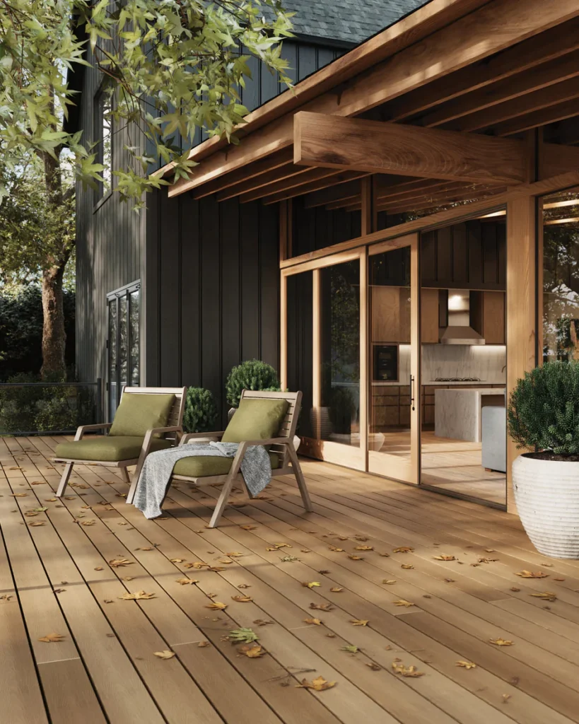 A backyard deck with light brown composite boards, green cushioned chairs, and fallen autumn leaves, set against a modern home with large glass doors.