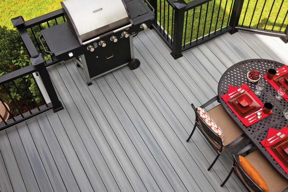 A grilling and dining setup on gray composite decking, featuring a black metal table with red dishware and a stainless steel barbecue grill.