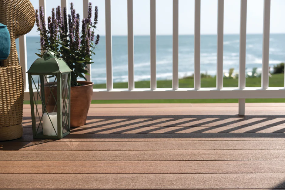 A close-up of medium brown composite decking on a coastal balcony with potted plants, a lantern, and an ocean view in the background.