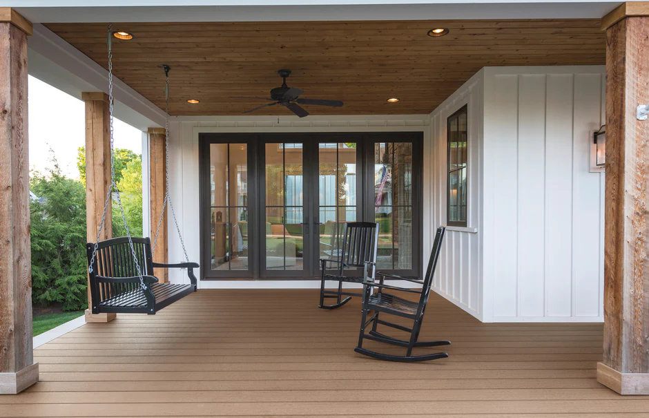 Charming farmhouse-style porch featuring Promenade decking in Sandy Pier, with natural wood accents, a black porch swing, and cozy rocking chairs for a timeless outdoor retreat.