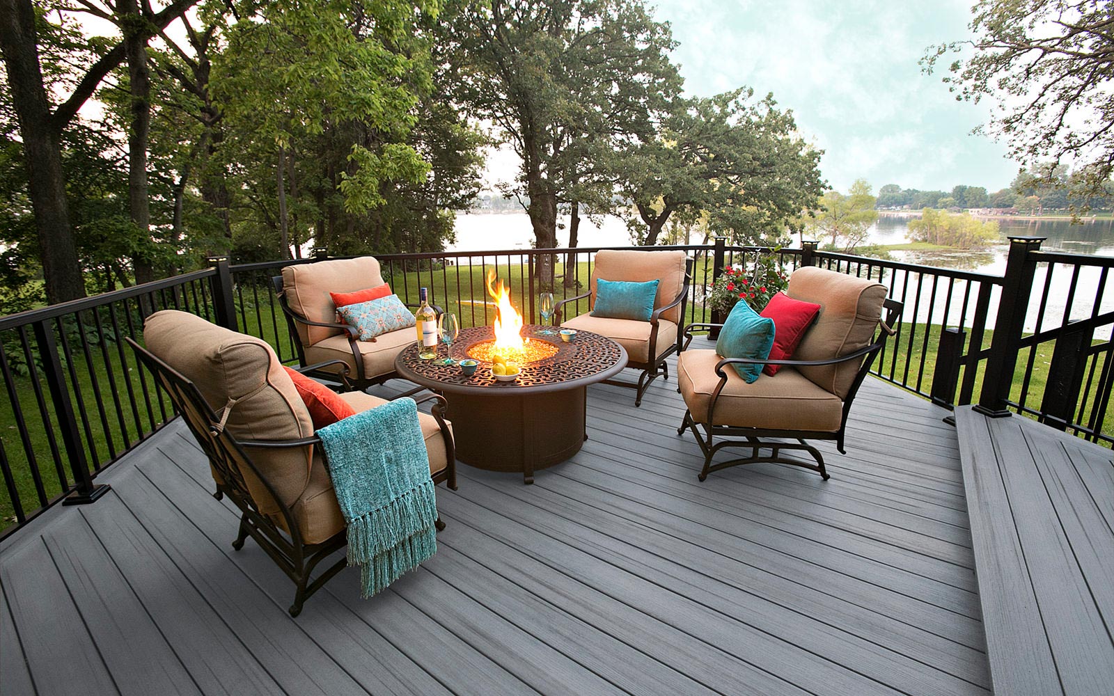 Light gray composite decking surrounding a fire pit with cushioned seating, black railings, and a scenic wooded lake backdrop.