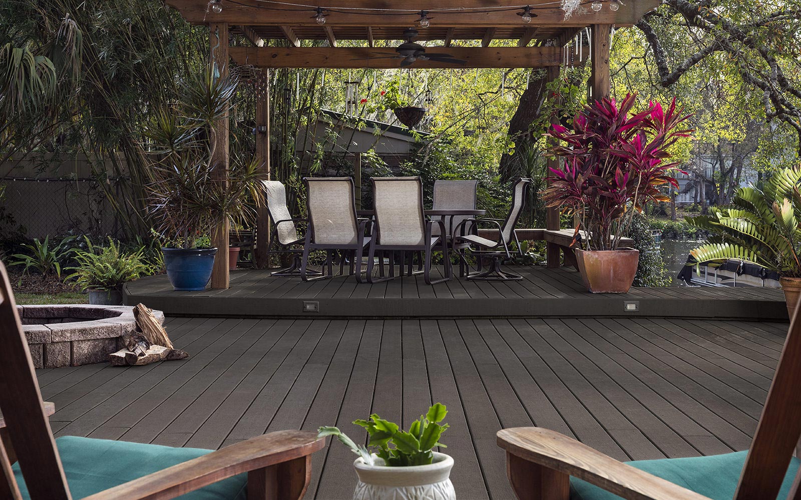 Weathered grey composite decking with a wood-like finish, featured under a pergola with natural furniture, tropical plants, and a stone fire pit.