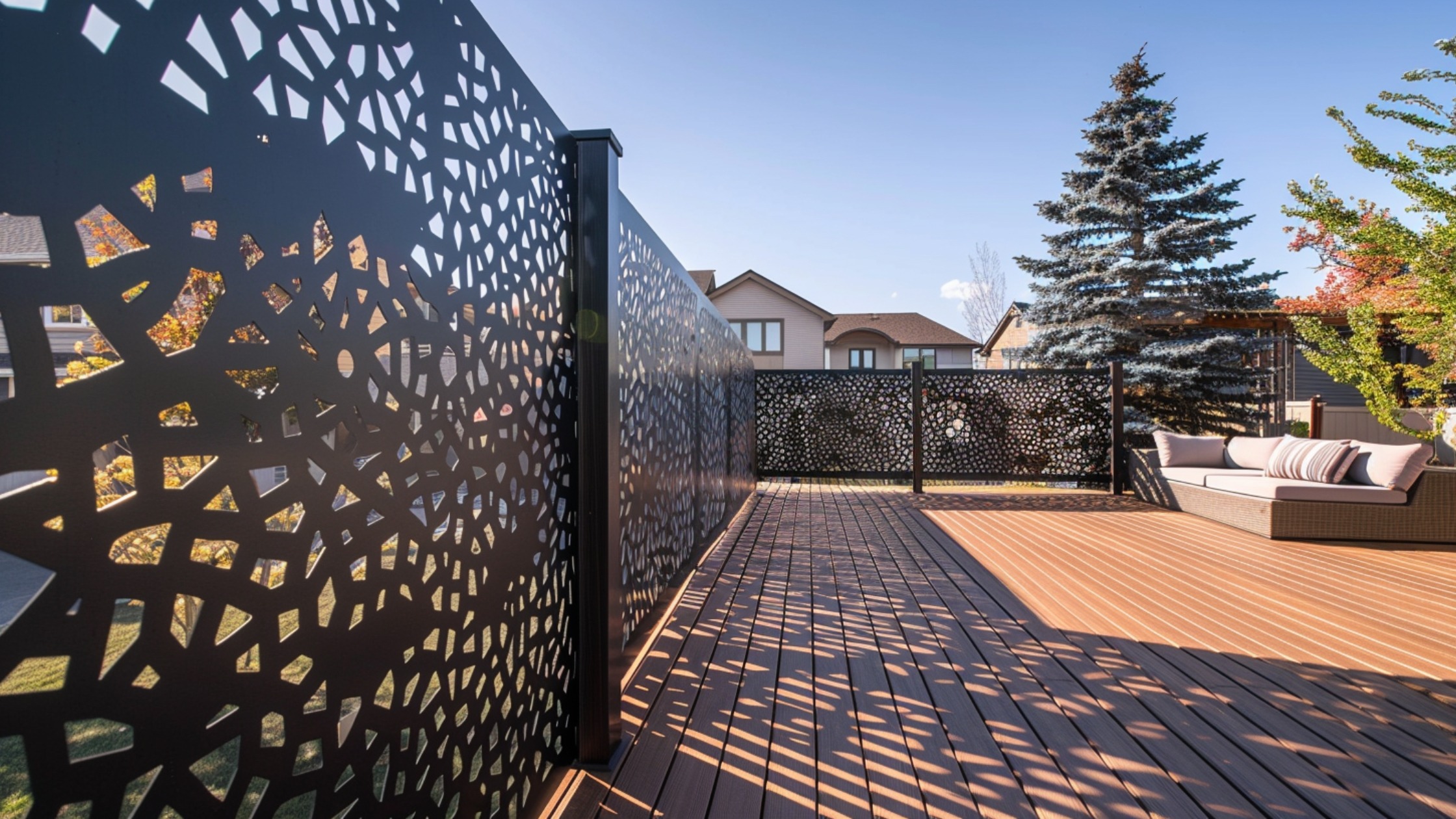 A large backyard deck featuring a tall, metal privacy screen with intricate cut-out patterns. The design casts artistic shadows across the space while blocking direct views from neighboring homes. The decorative panel serves as both a functional barrier and an eye-catching statement piece.