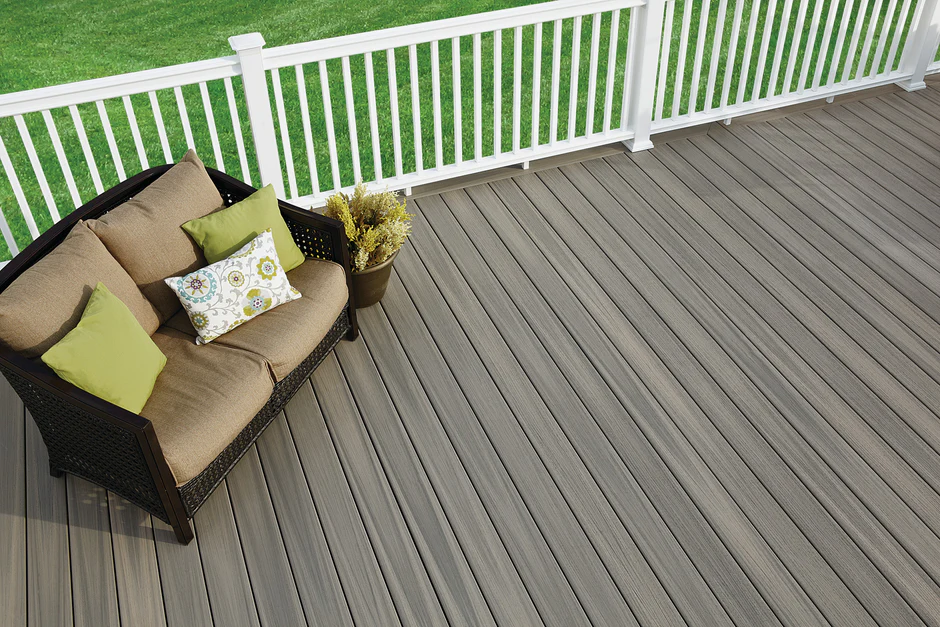 Deck built with Fiberon Paramount Sandstone decking featuring warm taupe-gray boards with subtle color variation. The outdoor space is styled with a cushioned wicker loveseat and green accent pillows, set against a white railing and bright green grass, showcasing the deck's modern and welcoming aesthetic.