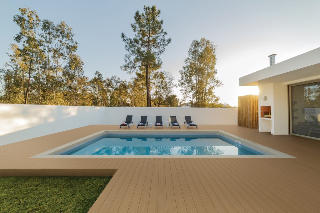 A backyard poolside deck built with Fiberon Paramount Mantel Clay decking boards surrounds a rectangular in-ground pool. The tan composite boards create a warm, cohesive look against the crisp white walls and modern home exterior. Five black lounge chairs line the back edge, and the sunlight casts soft shadows across the surface.