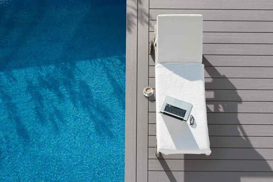 Fiberon Paramount Mineral decking installed poolside with a crisp, modern layout. The soft gray planks contrast against the bright blue water, with a white cushioned lounge chair, laptop, and coffee cup suggesting a relaxing outdoor workspace.