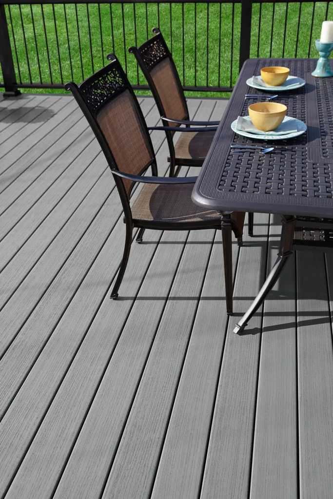 Outdoor dining area set on Fiberon Paramount Mineral decking. The deck features light gray composite boards with realistic grain texture, paired with black railing and dark metal dining furniture, creating a sleek, modern backyard setting.