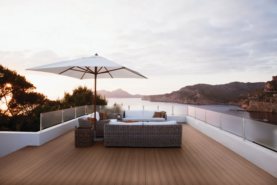 Rooftop outdoor space featuring Fiberon Promenade Russet Dune decking paired with a wicker sectional and umbrella, all set against a scenic mountain and water view at sunset. The brown decking blends seamlessly with the natural surroundings, creating a cozy and luxurious retreat.