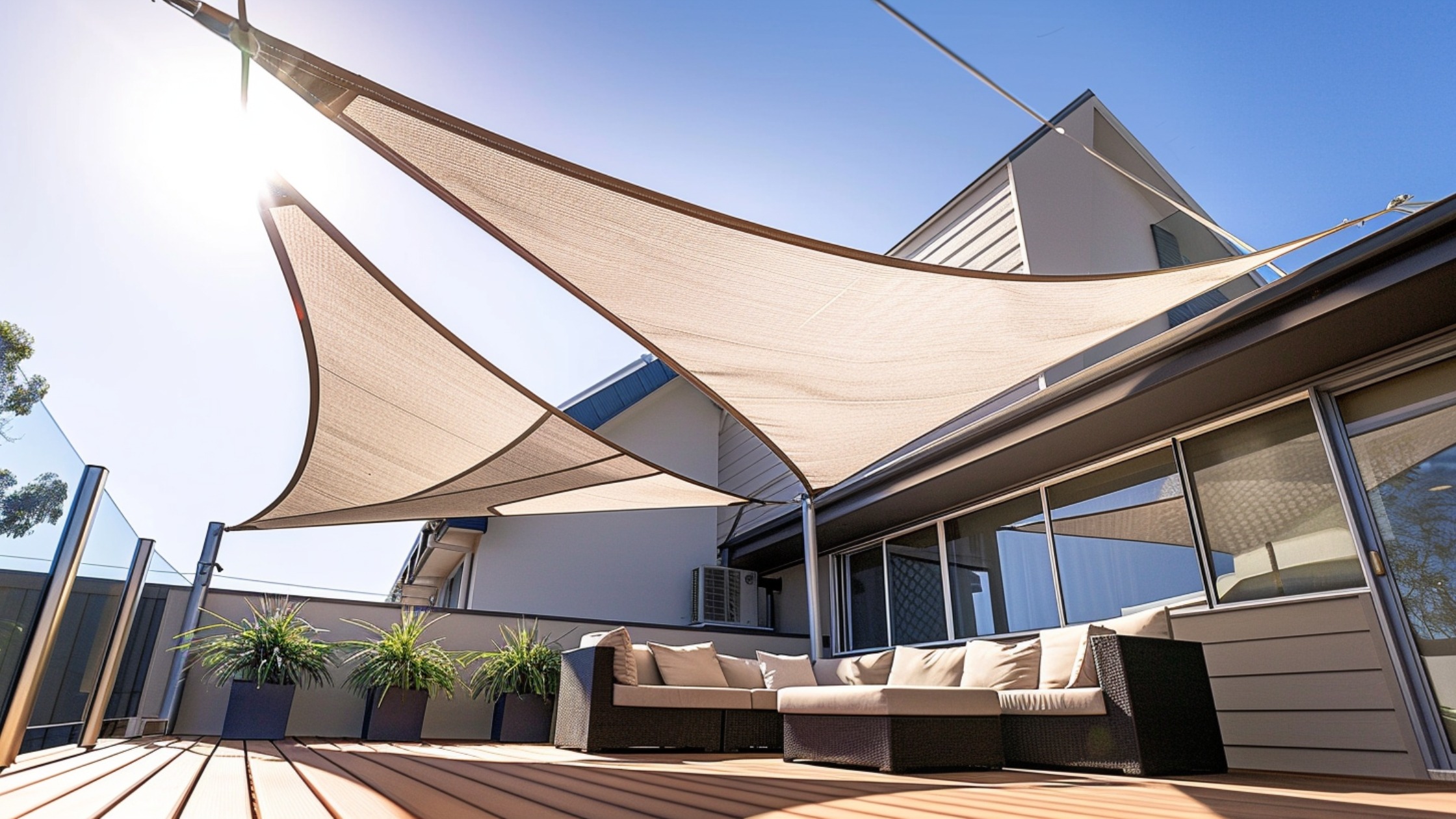 A backyard deck covered with large, triangular shade sails in a soft beige color. The angled design blocks views from above and provides sun protection. The sails stretch between posts and the house, giving the deck a modern, airy feel while creating a semi-private environment.