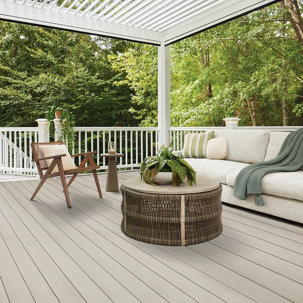 A serene outdoor lounge showcases TimberTech Harvest+ Timber Gray decking beneath a modern white pergola. The deck boards display a soft, cool-toned gray with subtle wood-like graining that mimics painted pine or gently weathered driftwood. Styled with a natural woven coffee table, modern wood-frame chair, and a cozy sectional layered with sage and cream accents, the space feels calm and elevated. The cool gray PVC decking balances beautifully against the surrounding lush greenery and crisp white railing.