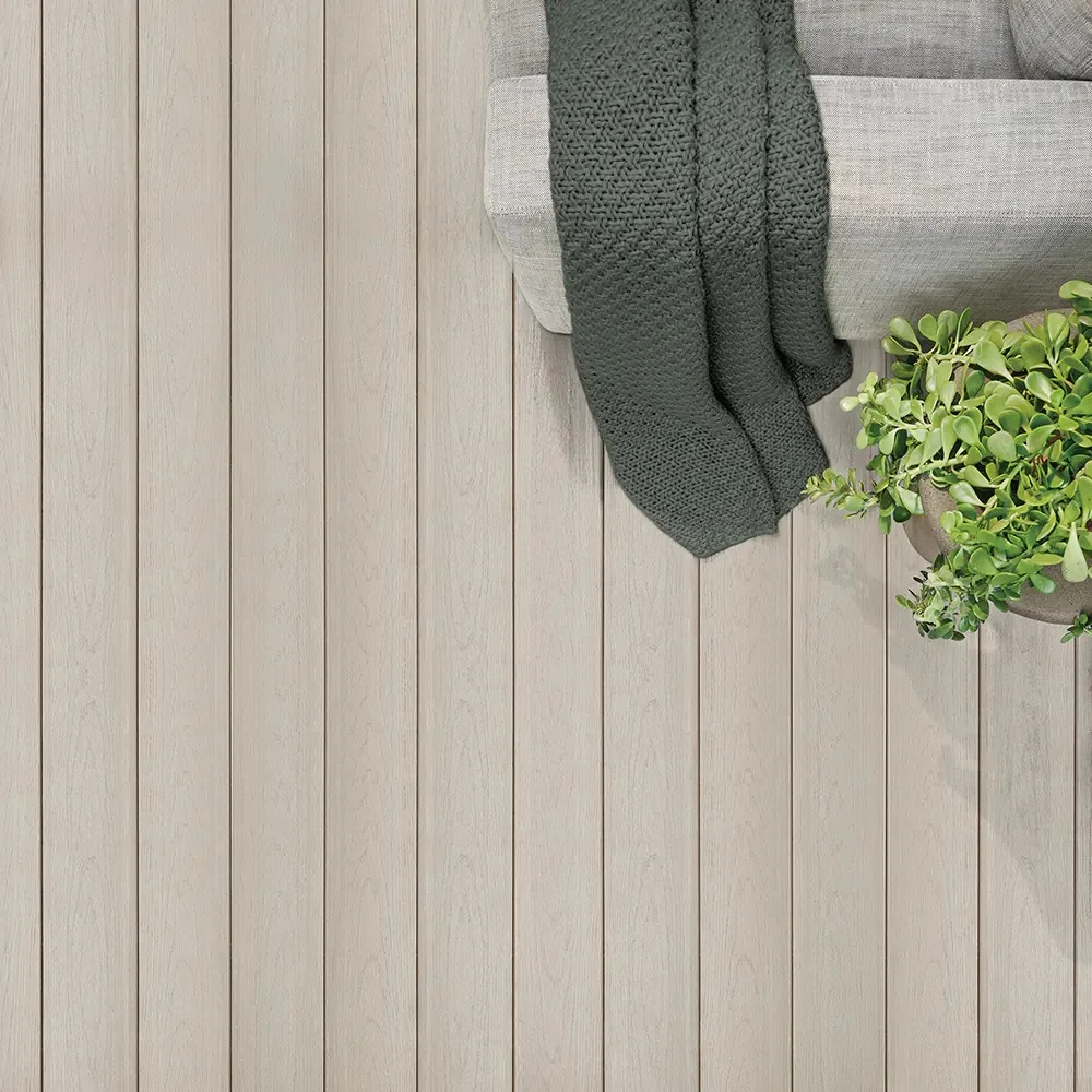 An overhead view of TimberTech Harvest+ Timber Gray decking, where soft, muted gray boards stretch across the frame with low-contrast woodgrain detailing. The tone resembles bleached pine or misty silver birch, delivering a clean, modern look. A sage green knit throw drapes over a neutral-toned sofa in the corner, while a lush green plant adds a fresh pop of color. The advanced PVC decking offers a matte finish and stays cool underfoot, ideal for relaxing outdoor moments.