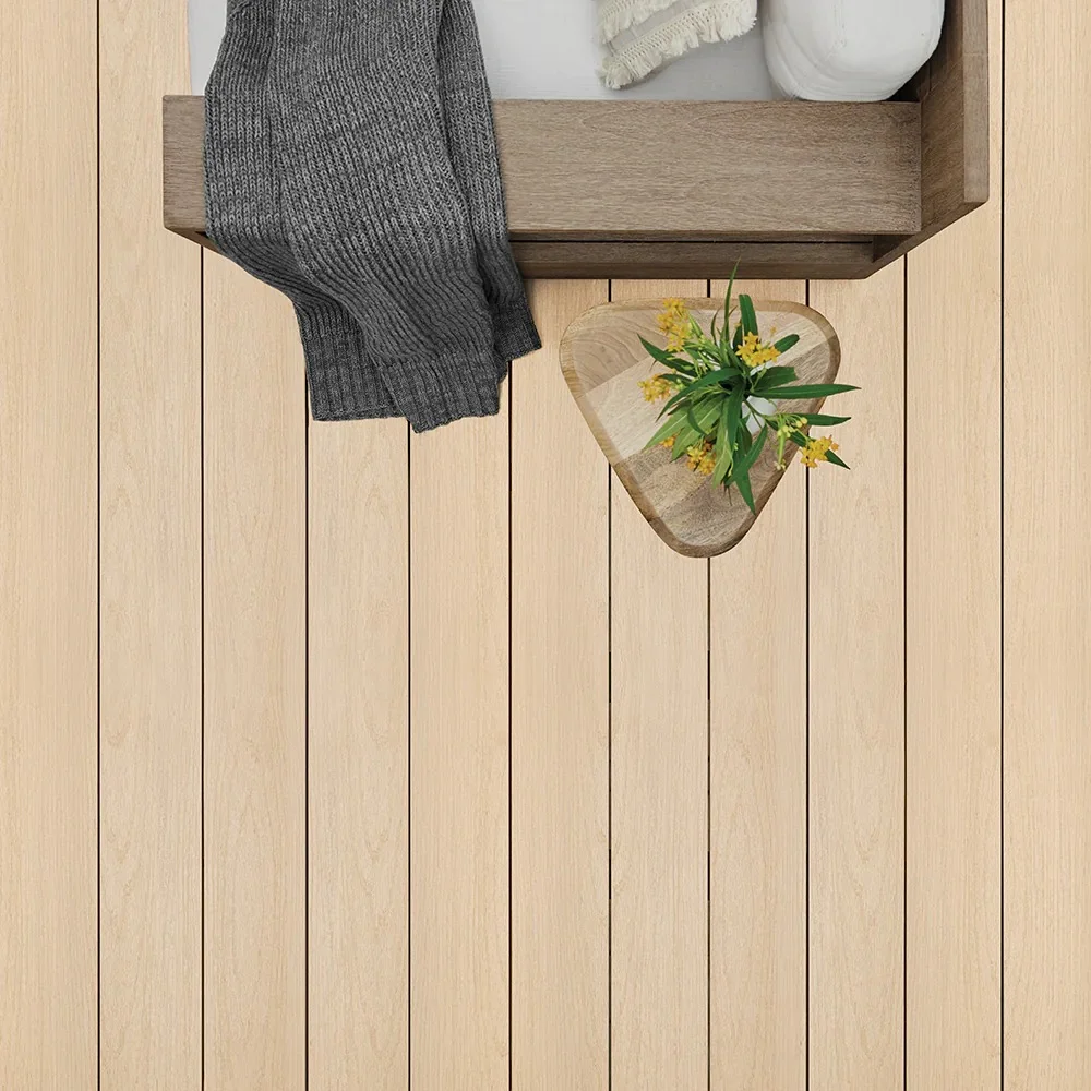 Close-up overhead view of TimberTech Harvest+ Toasted Wheat decking boards in a clean, light wood tone. The image includes a cozy corner of a cushioned seat, a gray knit blanket, and a rustic triangle side table holding a small bouquet of yellow flowers.
