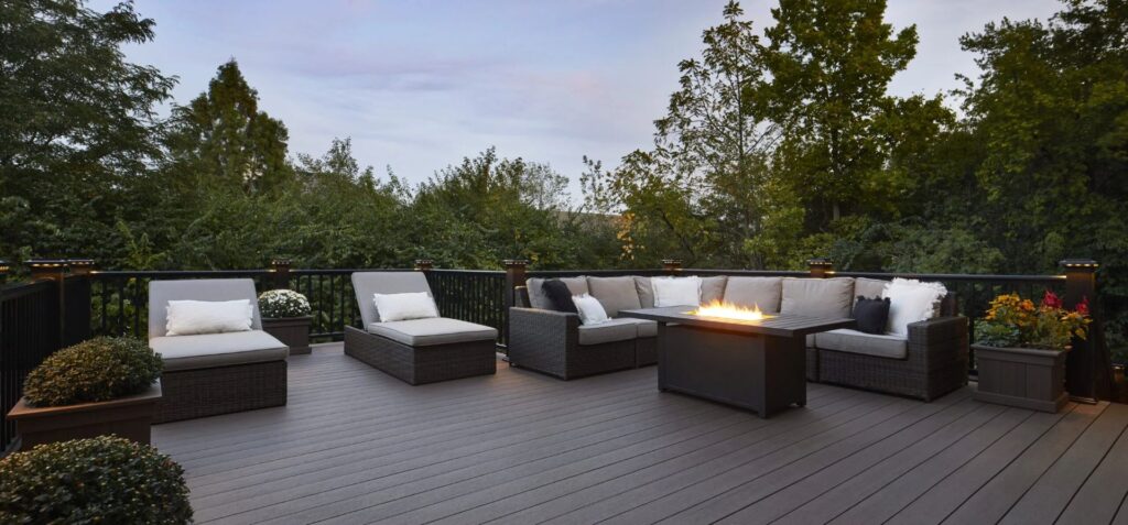 Backyard deck featuring TimberTech Landmark American Walnut decking, styled with a modern outdoor sectional and fire pit surrounded by lush green trees.