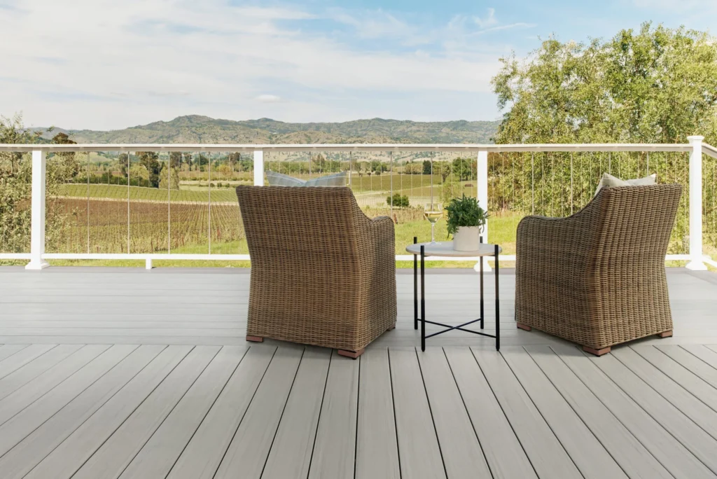 Two wicker chairs and a small round table are placed on a TimberTech Landmark Boardwalk deck overlooking a stunning vineyard and mountain view. The deck boards are a soft gray with subtle graining, and the white cable railing adds a sleek, open-air feel to the scene.