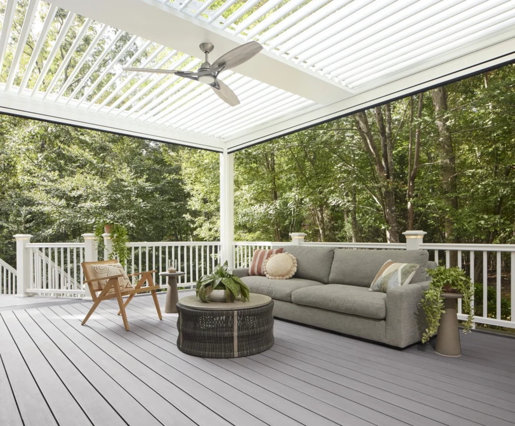 A cozy outdoor living space features TimberTech Landmark Boardwalk decking under a white pergola with a ceiling fan. A cushioned gray sofa, natural wood accent chairs, and leafy potted plants enhance the relaxing ambiance, with a forested backdrop adding privacy and shade.