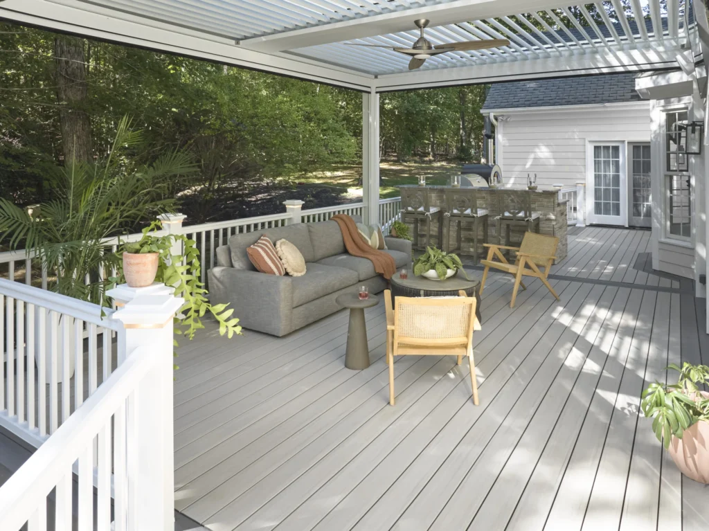 An expansive TimberTech Landmark Boardwalk deck is furnished with a sectional sofa, woven chairs, and multiple plant arrangements. A pergola provides partial shade, while the layout transitions to a built-in bar and grill area—ideal for hosting and entertaining outdoors.