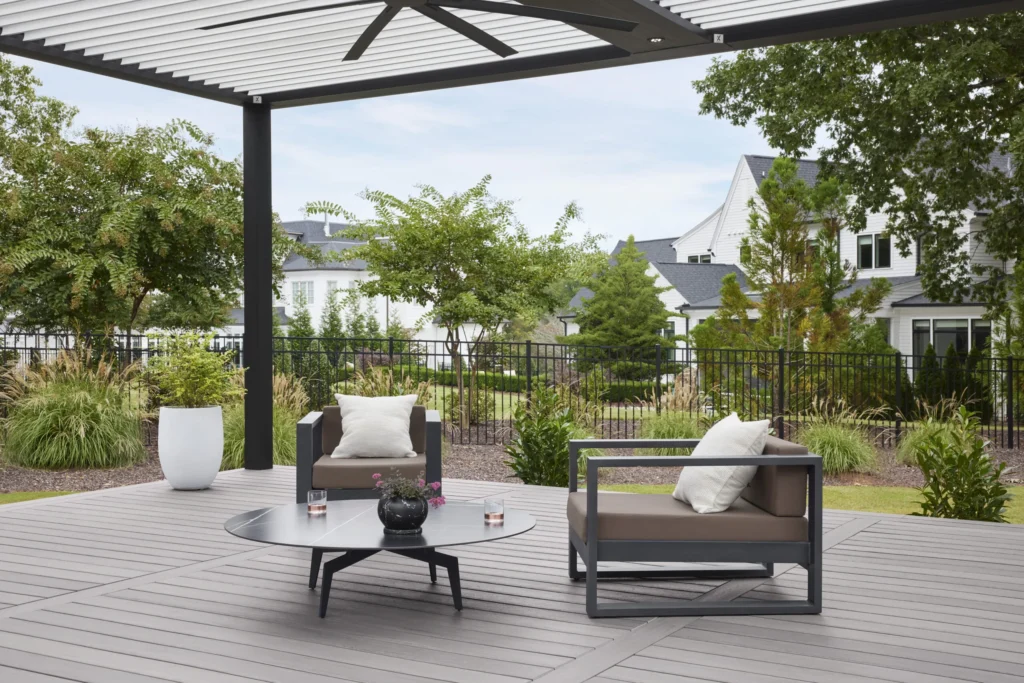 Modern outdoor lounge featuring TimberTech Landmark Collection decking in Castle Gate. The gray composite deck boards are arranged in a stylish pattern beneath a black pergola with a ceiling fan. Sleek gray patio furniture with white cushions and a low round coffee table add a sophisticated touch. The landscaped backyard includes ornamental grasses, shrubs, and a black metal fence enclosing a suburban neighborhood.