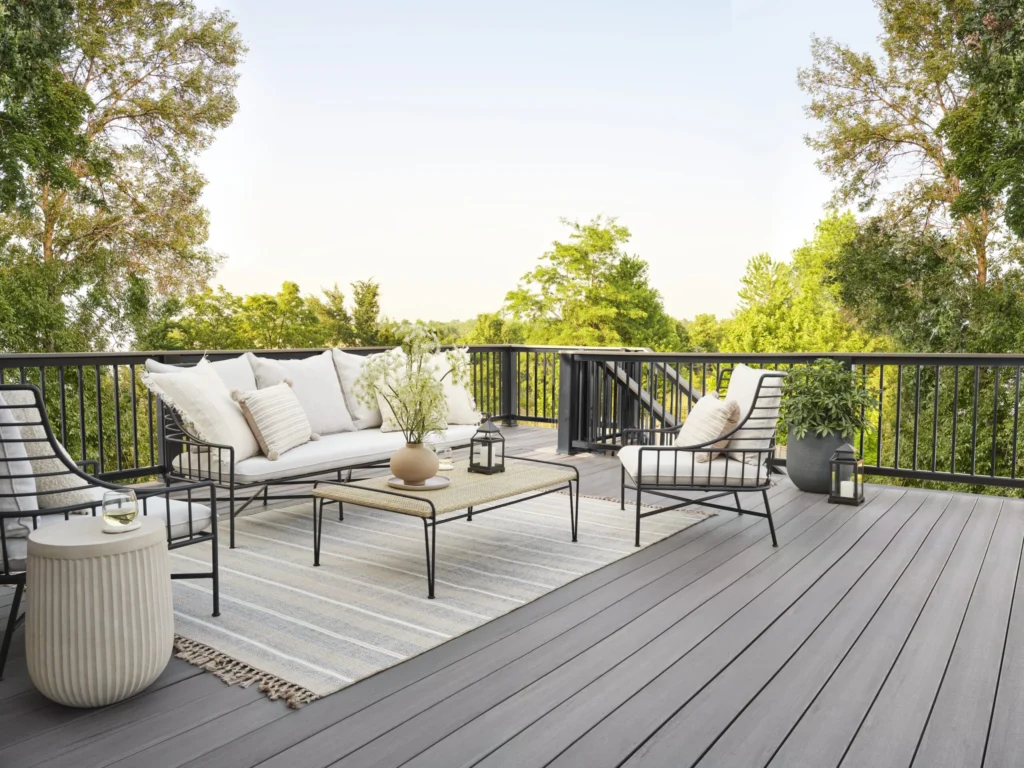 Spacious backyard deck built with TimberTech Castle Gate decking from the Landmark Collection. The gray composite boards contrast beautifully with a black railing and minimalist outdoor furniture. A striped rug, cream-colored cushions, and a neutral-toned coffee table create an airy, relaxing vibe. Lush green trees and bright sky complete the tranquil outdoor scene.