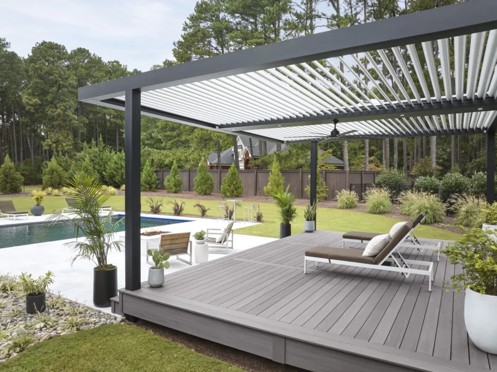 Poolside oasis featuring TimberTech Castle Gate composite decking from the Landmark Collection. A modern pergola with white louvers and black frame shades the gray deck. Two sleek chaise lounges with brown cushions invite relaxation. Potted plants and manicured landscaping surround the pool and lawn, creating a luxurious resort-style atmosphere.