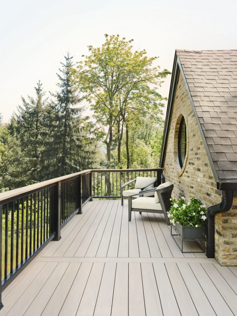 An elevated deck features TimberTech Landmark French White Oak boards with a natural woodgrain look, bordered by sleek black railings. The space includes two cushioned chairs and a planter, offering a quiet and sophisticated seating area overlooking tall pine trees and a peaceful yard.