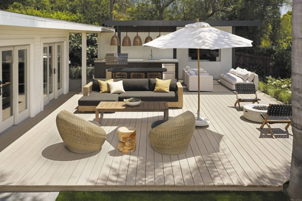 A modern backyard setup includes a large TimberTech Landmark French White Oak deck with a smooth, sun-washed finish. The area is furnished with a dark outdoor sectional, wicker chairs, a firepit, and a white patio umbrella, all nestled in a lush landscape with a built-in kitchen in the background.