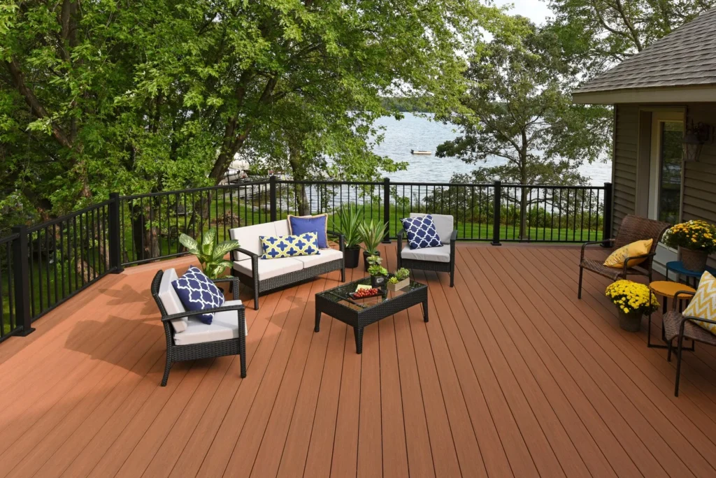Spacious lakeside deck built with TimberTech Vintage Cypress boards, featuring a reddish-brown tone, black railing, and patio furniture with blue and yellow cushions, surrounded by trees and lake views.