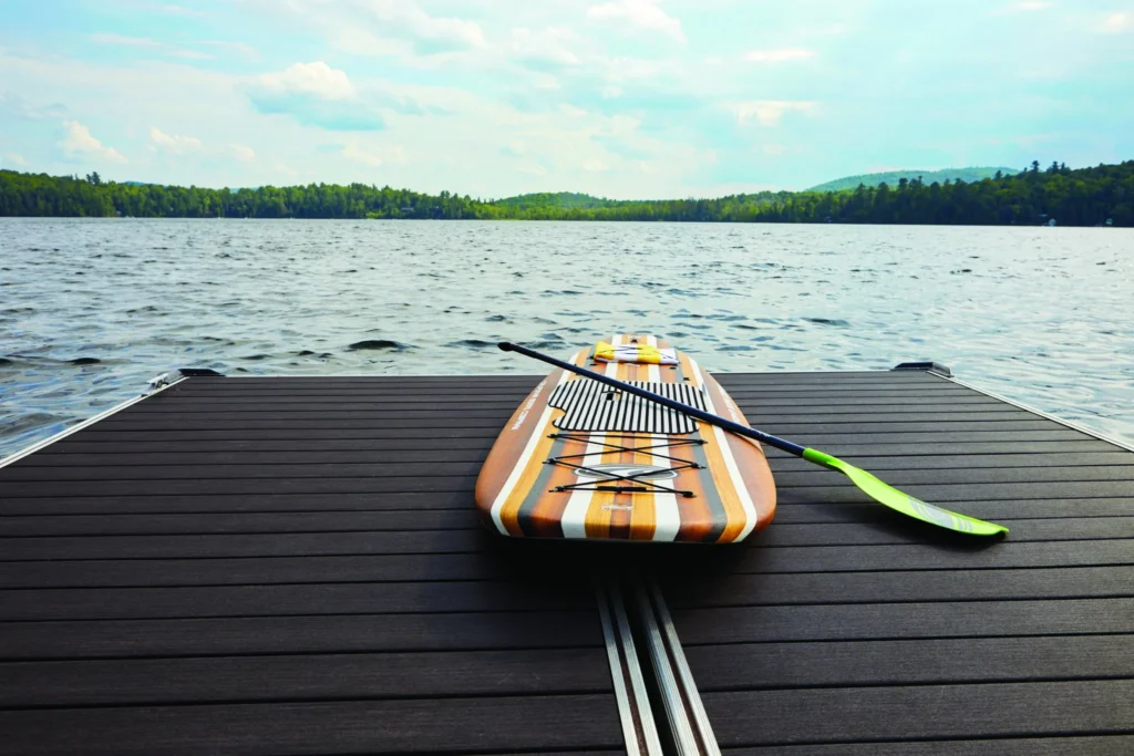 A striped paddleboard rests on TimberTech Vintage Dark Hickory decking at the edge of a serene lake, with forested hills in the background.