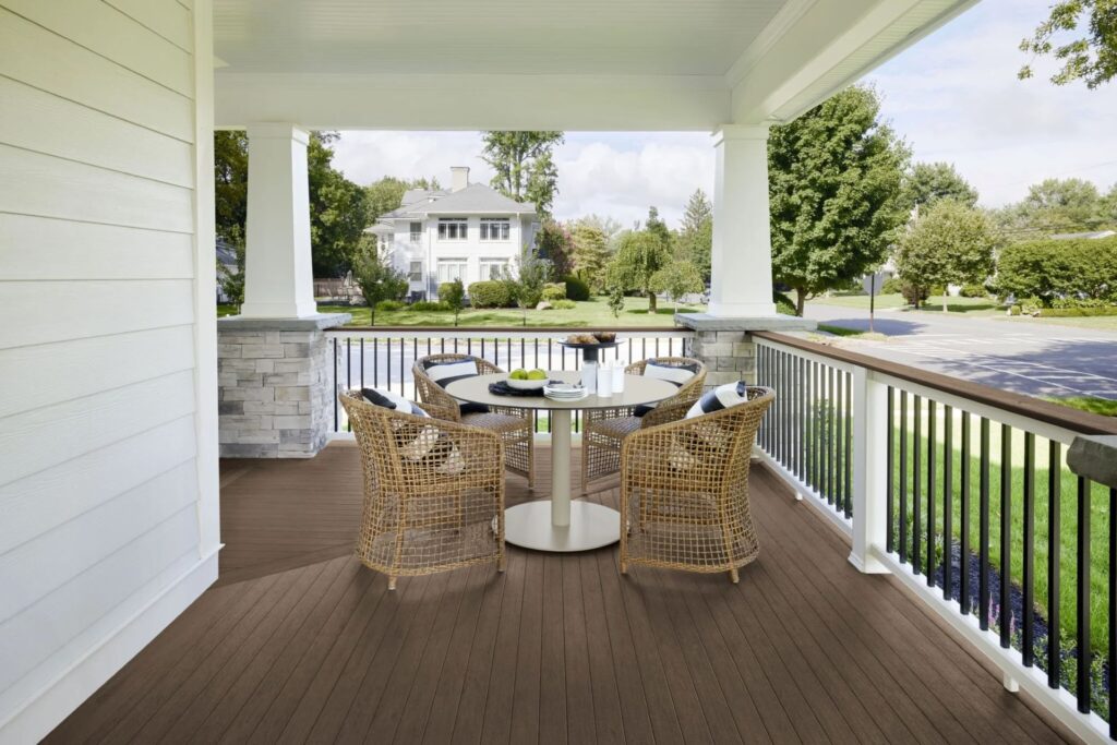A covered front porch features TimberTech Vintage English Walnut Advanced PVC decking with rich brown tones and natural wood grain texture. The space includes a round cream-colored table surrounded by four woven rattan chairs with black and white pillows. White pillars, stone bases, and a white-and-black railing add classic charm, while the surrounding neighborhood with lush greenery completes the peaceful, upscale setting.
