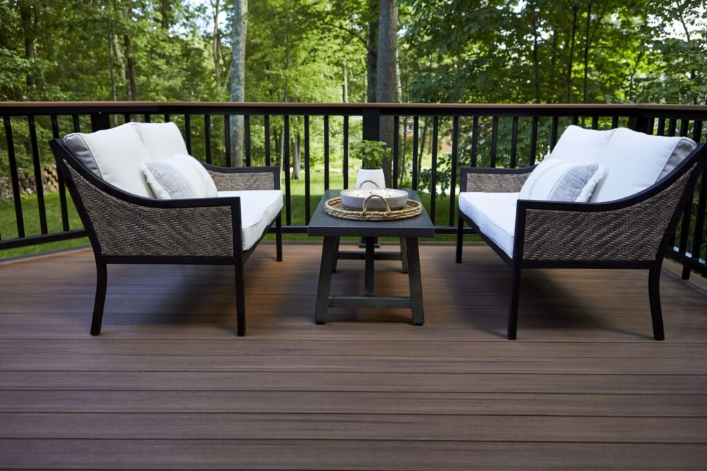 Outdoor seating area on a deck built with TimberTech Vintage Mahogany decking boards, featuring two cushioned loveseats and a coffee table with woven details, surrounded by lush green trees and a black railing.