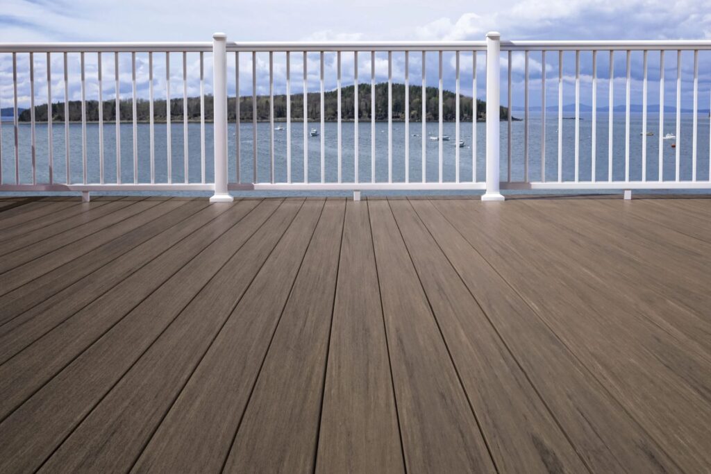 Close-up view of TimberTech Vintage Mahogany decking boards on a waterfront deck with white railings, overlooking a serene lake and forested island.