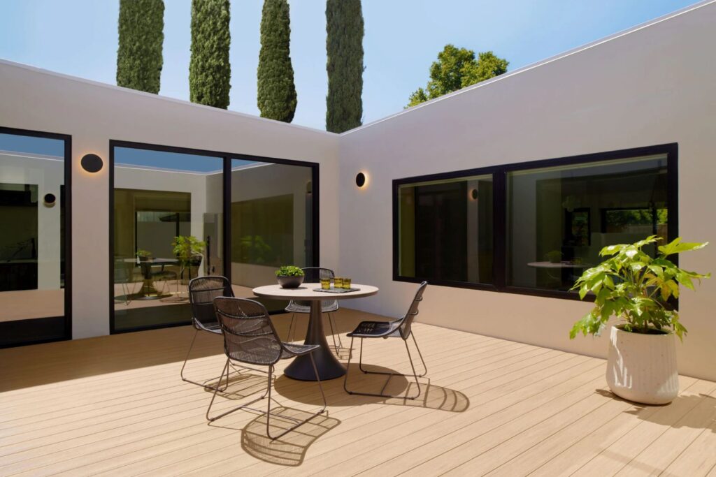 TimberTech Vintage Weathered Teak deck in a minimalist courtyard, featuring a modern round dining table, black mesh chairs, and bold white architecture with large glass windows.