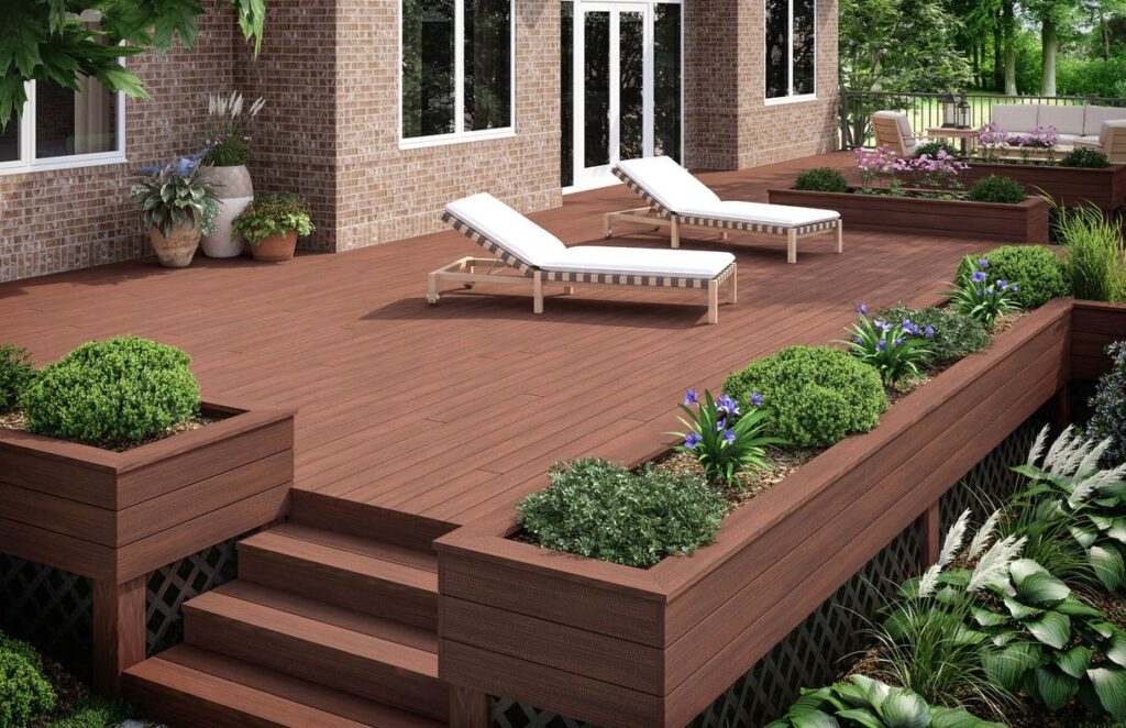 Spacious backyard deck built with Deckorators Distressed Decking in Cherrywood, featuring deep reddish-brown tones with a warm, inviting woodgrain texture. Two cushioned lounge chairs rest on the main deck, while raised planters with lush greenery and purple flowers line the perimeter. The cherrywood planks have a low-gloss finish that mimics real hardwood and pair beautifully with the surrounding garden and brick home exterior.