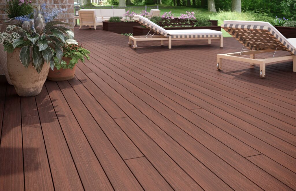 Close-up view of Deckorators Distressed Decking in Cherrywood, showcasing its realistic woodgrain texture and rich, dark red-brown coloring. The deck surface surrounds two lounge chairs with light cushions and checkered patterns. Visible wood detail mimics authentic cherry hardwood, creating a cozy, upscale outdoor living space with vibrant planters and surrounding greenery in the background.