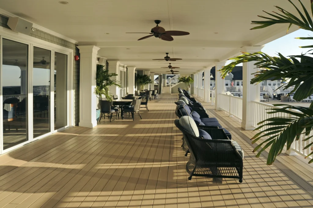Covered veranda featuring TimberTech Harvest Brownstone advanced PVC decking boards in a warm, medium brown tone. The scene shows wicker lounge chairs with navy cushions and ceiling fans overhead, evoking a coastal resort feel. The durable PVC decking boards run lengthwise across the entire space, highlighting a uniform, low-maintenance surface ideal for hospitality settings.