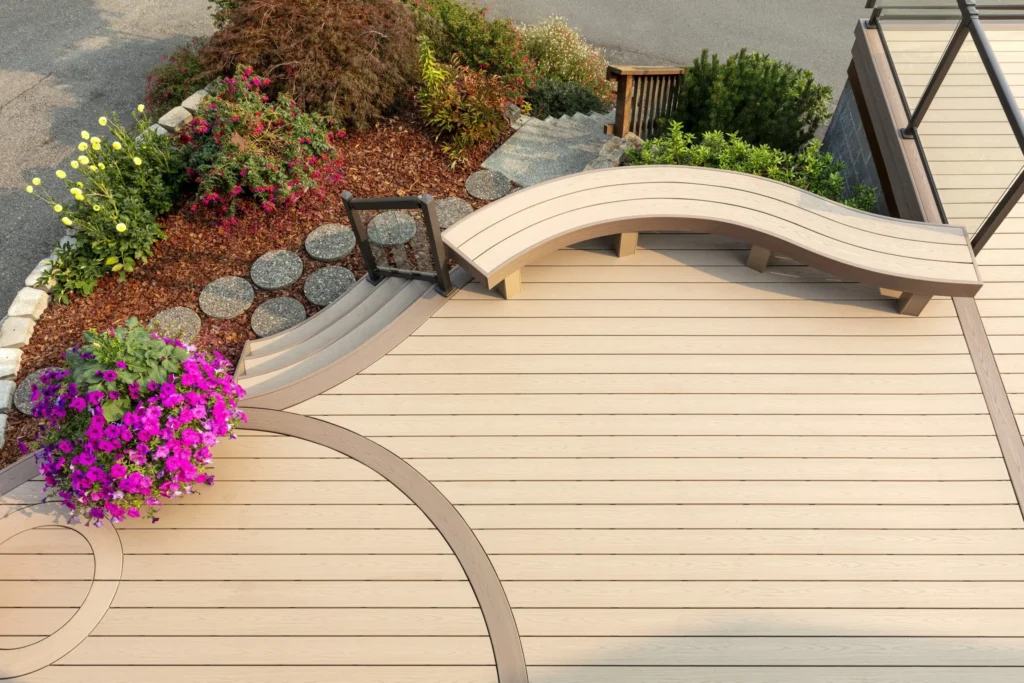 Beautifully curved backyard deck built with TimberTech Harvest Brownstone advanced PVC decking in a light beige finish. The space includes a built-in curved bench, circular inlay patterns in contrasting tones, and steps leading to a landscaped garden with vibrant flowers. The composite PVC decking offers long-lasting beauty with natural wood-like grain and superior resistance to moisture and fading.