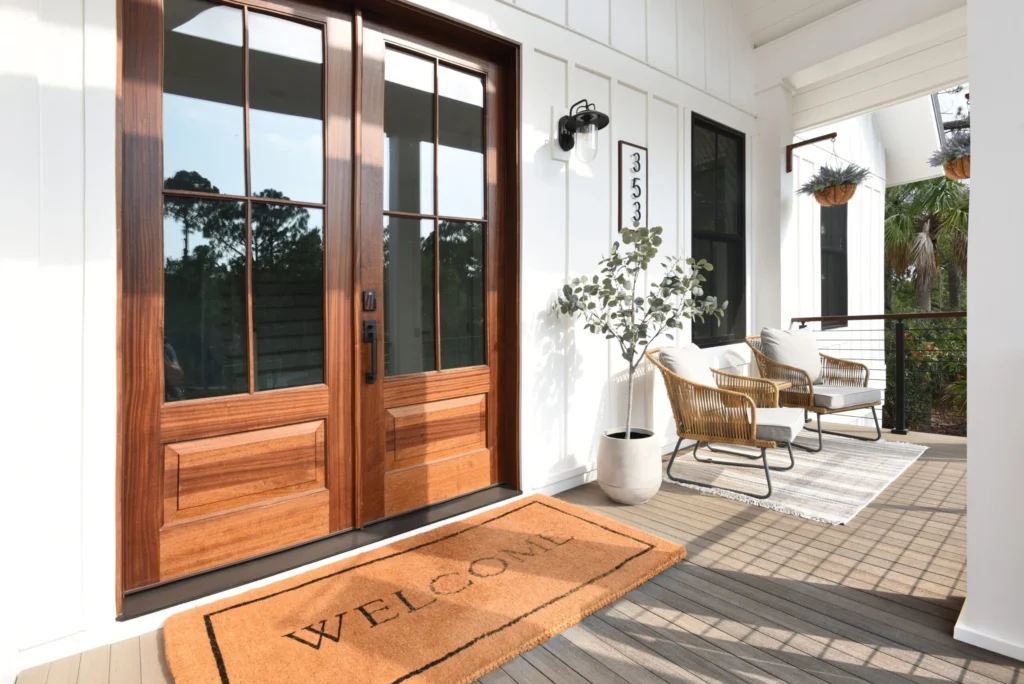 A welcoming front porch with TimberTech Porch Coastline boards, showcasing subtle gray-brown hues beneath a warm wooden door. Two wicker lounge chairs with cushions and a “WELCOME” mat add charm to this bright, farmhouse-style entryway.