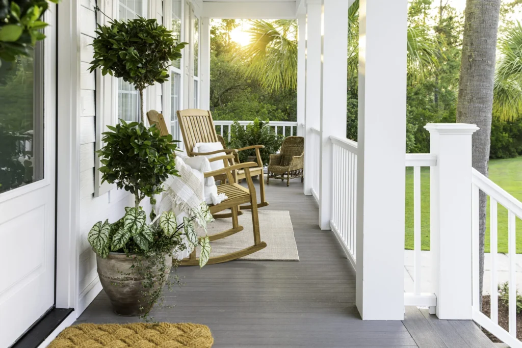A classic southern-style wraparound porch featuring TimberTech Porch Coastline boards in a weathered taupe shade, paired with natural wood rocking chairs and lush greenery under a white railing.