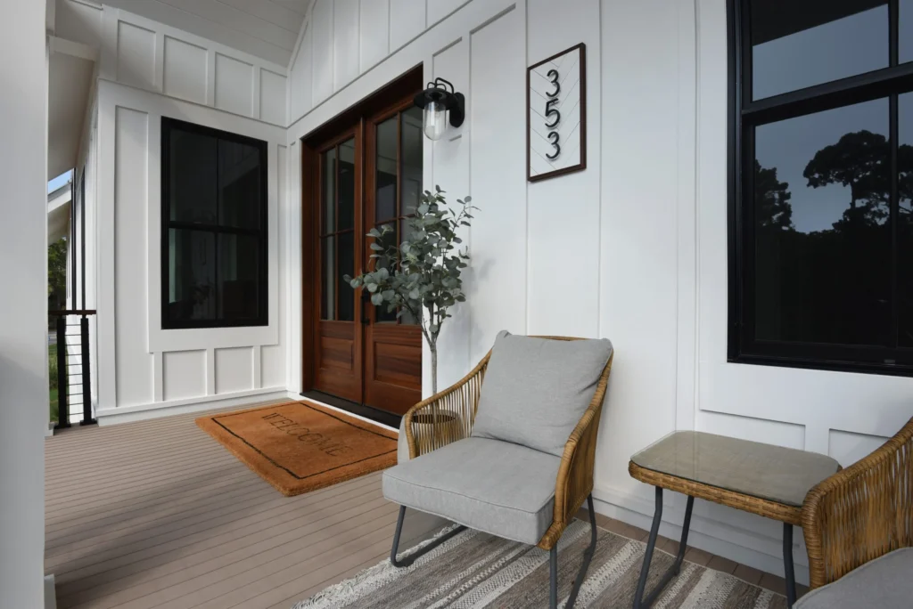TimberTech Porch Coastline boards under a shaded, modern farmhouse porch with clean white siding, dark-framed windows, and a soft neutral palette. Wicker seating and a woven rug complement the light gray wood tones of the porch floor.