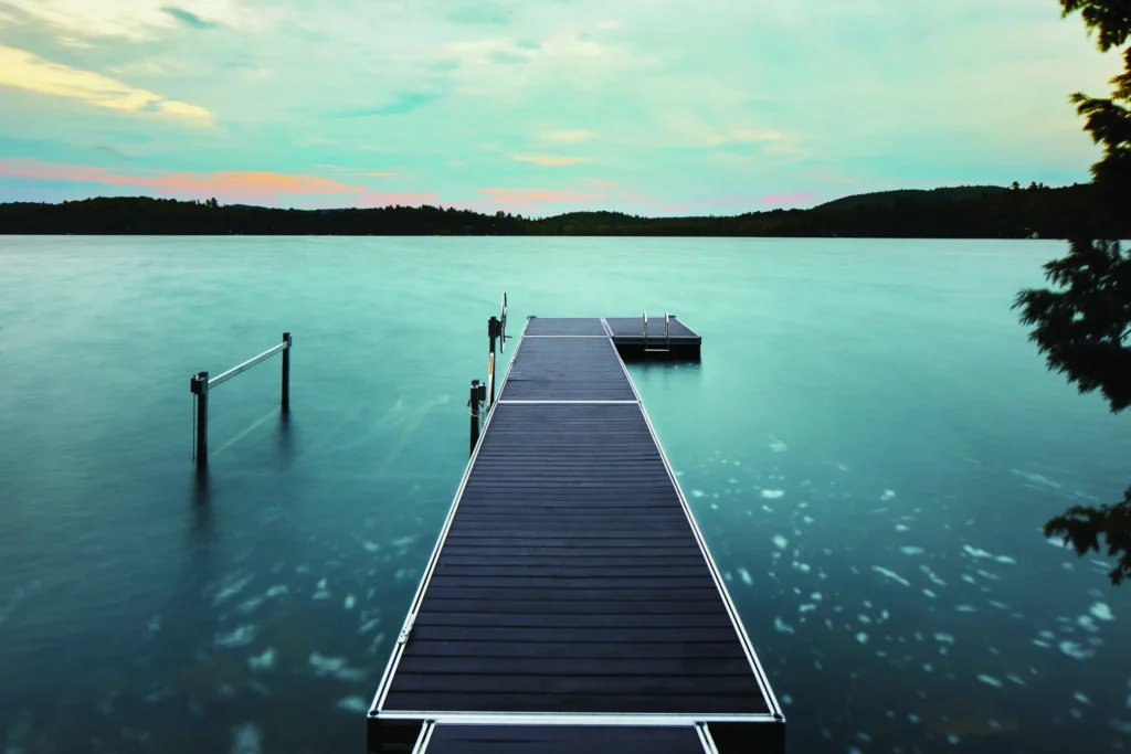 A long dock made with TimberTech Porch Dark Hickory Advanced PVC porch boards extends over a calm lake at sunset. The rich, charcoal-toned decking features a clean tongue-and-groove layout, bordered by sleek aluminum framing, creating a serene and sophisticated waterfront retreat.
