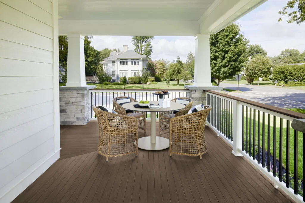 Covered front porch with white pillars and railing, featuring TimberTech Porch decking in English Walnut, a rich brown tone with natural woodgrain detail, paired with wicker furniture and a round table in a suburban neighborhood.